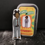 Mini fabric doll of artist Jean-Michel Basquiat beside a keepsake gift tin. All set against a dark background.