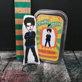 Robert Smith mini fabric doll next to a gift tin. set against a black and textured background.