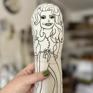 Black and white illustrated fabric doll of singer Dolly Parton held up by a hand.