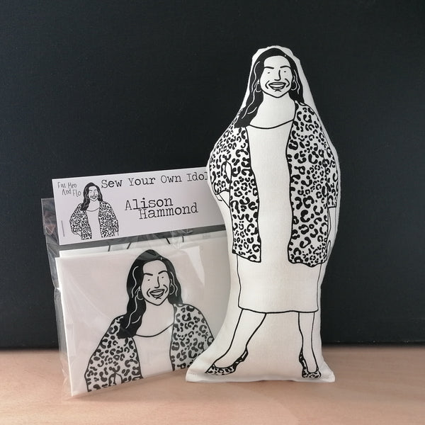 Sewing craft kit and black and white screen printed fabric doll cushion featuring The Great British Bake Off and This Morning host, Alison Hammond. Stands against a black background on a light wood table.