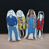 Four mini colourful fabric dolls featuring illustrations of The Great British Bake Off hosts and judges, Paul Hollywood, Prue Leith, Alison Hammond and Noel Fielding. Stands against a black background on a light wood table.