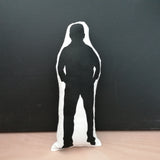 Reverse of a black and white screen printed fabric doll cushion featuring a silhouette of The Great British Bake Off and judge, Paul Hollywood. Stands against a black background on a light wood table.