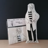 Craft kit and screen printed fabric doll cushion featuring The Great British Bake Off judge, Prue Leith. Stands against a black background on a light wood table.