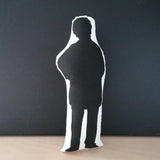 Reverse of a black and white screen printed fabric doll cushion featuring a silhouette of The Great British Bake Off and judge, Prue Leith. Stands against a black background on a light wood table.