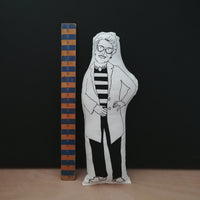 Black and white screen printed fabric doll cushion featuring The Great British Bake Off and judge, Prue Leith. Stands against a black background on a light wood table. To the left is a wooden ruler.