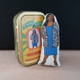 Keepsake tin and mini colourful fabric doll featuring Bake Off and This Morning host, Alison Hammond. Stands against a black background on a light wood table.