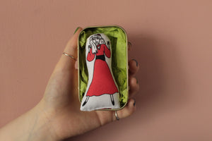 Mini fabric doll of singer Kate Bush in a tin. This is held in an open hand against a pink wall.