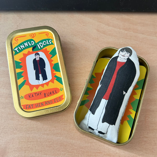 Mini fabric collectible doll and keepsake tin featuring an illustration of actor and comedian, Kathy Burke. On a wooden surface.