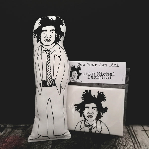 Black and white fabric doll of Jean-Michel Basquiat beside a craft kit and set against a black wall.