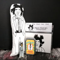 A selection of fabric dolls and craft kit featuring artist Basquiat. Stood against a black wall.