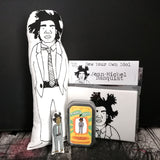 A selection of fabric dolls and craft kit featuring artist Basquiat. Stood against a black wall.