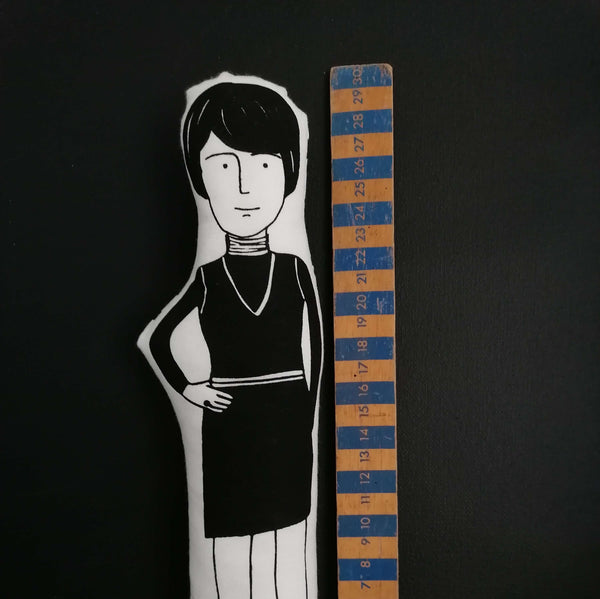 Black and white screenprinted fabric doll of British artist Bridget Riley beside a wooden ruler for scale.