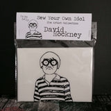 A sew your own doll craft kit of artist David Hockney against a black background.