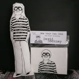 A black and white fabric doll of David Hockney alongside a craft kit of the same doll against a balck backgroound.