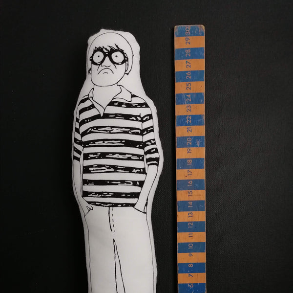 Black and white fabric doll of British artist David Hockney stood next to a wooden ruler for scale.