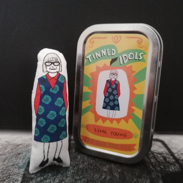 Mini fabric keepsake doll of  The Great British Sewing Bee host Esme Young beside a decorative tin with sticker detail. All against a black backdrop.