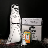 A selection of Freddie Mercury gifts including a monochrome fabric doll, a mini doll and tin and a craft kit, all against a black backdrop.