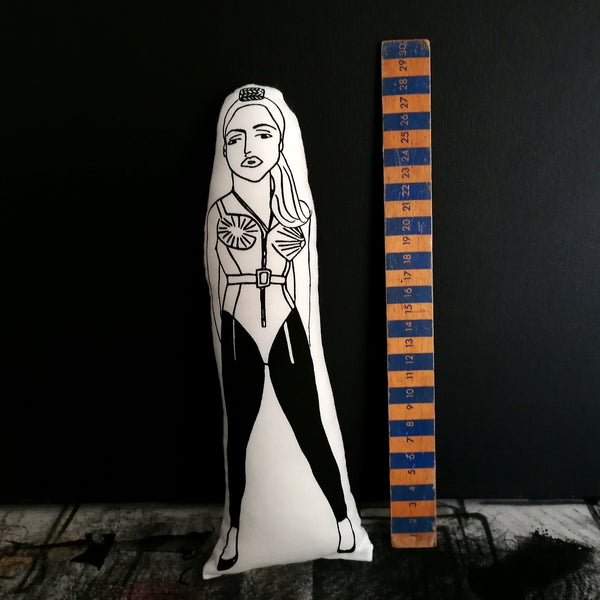 Black and white fabric doll featuring an illustrated design of Madonna. Beside a wooden ruler for scale and set against a black wall.
