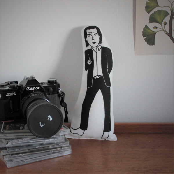 Black and white screen printed fabric doll of Nick Cave stood on a wooden table beside a pile of CDs and a Canon camera.