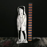 Screen printed black and white fabric doll of Noel Fielding. Stood beside a wooden ruler for scale against a black wall.
