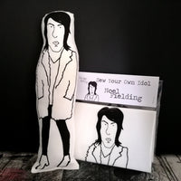 Black and white screen printed fabric doll of Noel Fielding beside a craft kit to sew your own Noel doll. All set against black painted backdrop.