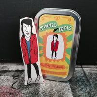 Mini fabric art doll of Noel Fielding next to a gift tin with sticker design of Noel. Set against a  black painted background.