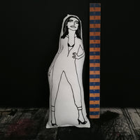 Black and white screen printed fabric doll of musician PJ Harvey in her iconic jumpsuit. Stood beside a wooden ruler to indicate the size.  All set against a black painted backdrop.