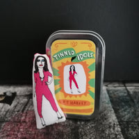 Mini fabric doll of PJ Harvey in her iconic pink jumpsuit beside a gift tin and set against a black backdrop.