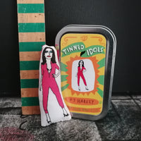 Mini fabric doll of musician PJ Harvey in her iconic pink jumpsuit next to a gift tin. They are both set against a black background and beside a vintage wooden ruler to show scale.