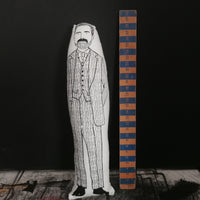 A black and white screen printed fabric doll  featuring an illustration of The Great British Sewing Bee host Patrick Grant. It is stood beside a wooden ruler against a black wall to indicate size.