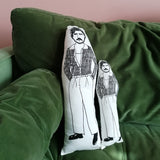 A green sofa against a pink wall. On the sofa are two black and white illustrated cushions featuring Pedro Pascal.
