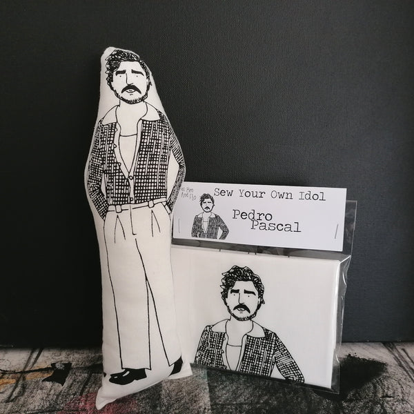 A black and white illustrated fabric doll of Pedro Pascal ans craft kit against a black wall.