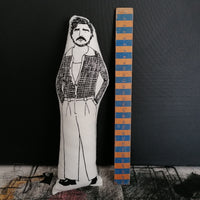 A black and white illustrated screen printed fabric doll of Pedro Pascal stood by a wooden ruler against a dark wall.