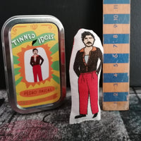 A small fabric doll of Pedro pascal beside a gift tin with sticker detail. Both stand before a black wall on a textured painted cloth. There is a wooden ruler beside the doll to show size of the doll which is 9cm high.