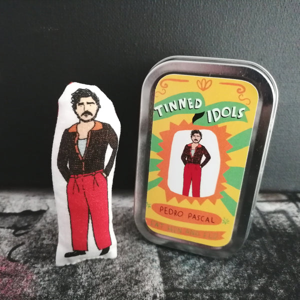A small fabric doll of Pedro pascal beside a gift tin with sticker detail. Both stand before a black wall on a textured painted cloth.