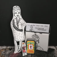 A selection of gifts of fashion designer Vivienne Westwood set against a dark backdrop. Includes a black and white screen printed fabric doll, a mini coloured fabric doll, a gift tin and a craft kit.