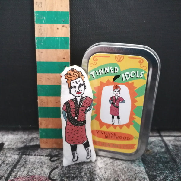 MIni fabric doll of Dame Vivienne Westwood between a vintage wooden ruler and a gift tin, set against a dark background. 