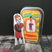 Mini fabric doll of Vivienne Westwood beside a gift tin with sticker design lid. All set against a dark backdrop.