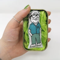 Mini fabric doll of Danny Devito in a tin held in a hand for scale against a white background.