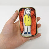 MIni doll of Greta Thunberg in a tin held in an open hand for scale.