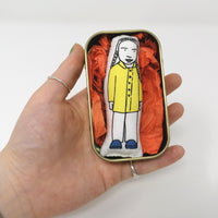 Mini doll of Greta Thunberg in a tin held in a hand.