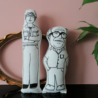 louis theroux and danny devito dolls on a shelf by a plant and empty gilt frame