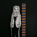 Close up of a black and white fabric doll of Artist Tracey Emin beside a wooden ruler against a black background.